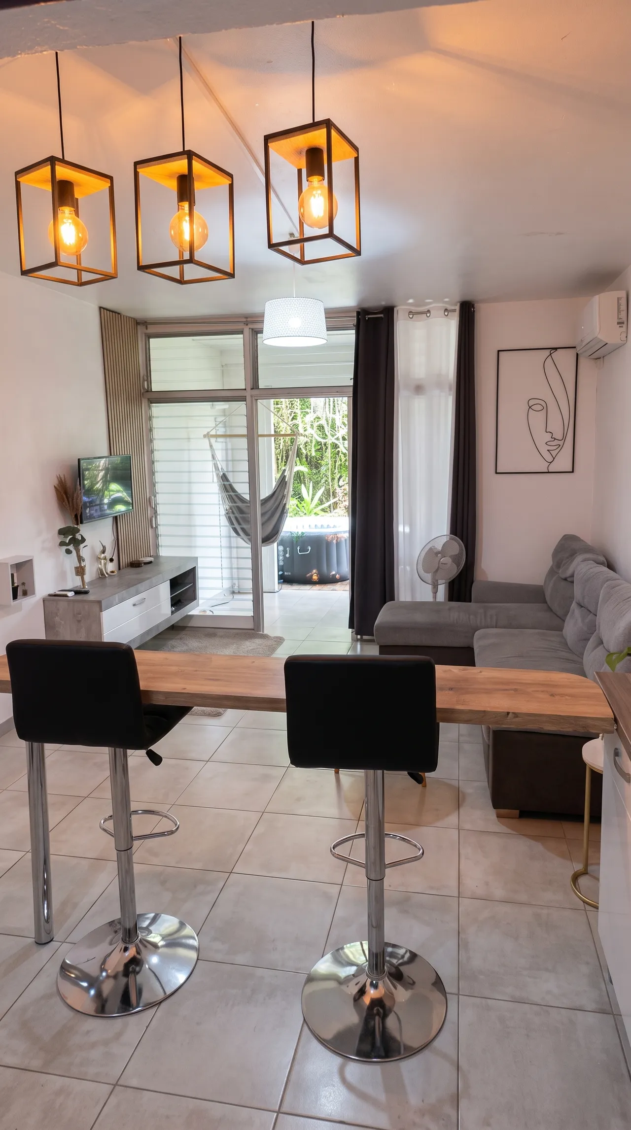 Cuisine de dos, vue sur la table de bar avec des chaises hautes noires, vue sur le lit et le salon TV